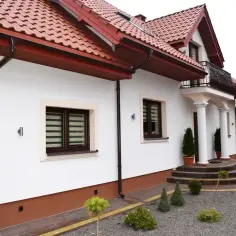 Biały dom z brązowym dachem i rynnami. Elegancki cokół podkreśla jego architekturę.