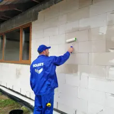 Pracownik w niebieskim kombinezonie nakłada wałkiem podkład na bloczki z betonu komórkowego. To kluczowy etap przygotowania pod tynk zewnętrzny.