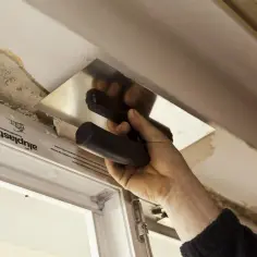 Ręka z kielnią wygładza tynk nad oknem. Widać fragmenty starego tynku i pianki montażowej.