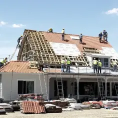 Ekipa budowlana pracuje nad wymianą dachu. Widać robotników na drabinach i rusztowaniach, a także nowe materiały budowlane.