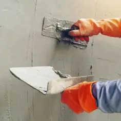 Ręce w pomarańczowych rękawiczkach nakładają tynk gipsowy na ścianę kielnią.