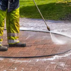 Osoba w zielonych spodniach z odblaskami myje kostkę brukową myjką ciśnieniową, usuwając stare plamy z oleju.