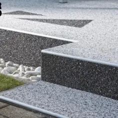 Nowoczesne schody z kamiennego dywanu, z metalowymi noskami, otoczone białymi kamieniami i zieloną trawą.