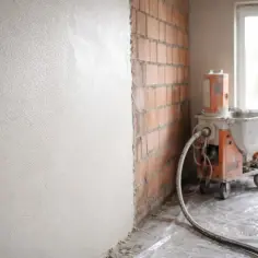 Maszyna do tynkowania w trakcie pracy. Ściana pokryta świeżym tynkiem, obok ceglana ściana i okno. Wybór między tynkami gipsowymi czy cementowo wapienne.