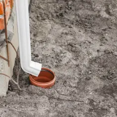 Biała rura spustowa odprowadza wodę z rynien do ceramicznego kręgu w ziemi.