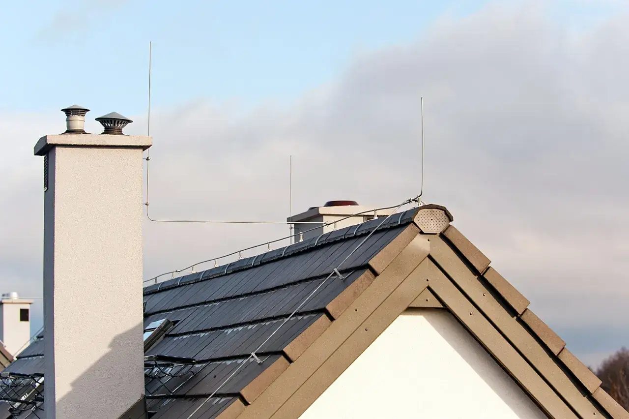 Dach z ciemną dachówką, komin i piorunochron. System ochrony przed wyładowaniami atmosferycznymi, czyli piorunochron, zabezpiecza budynek.