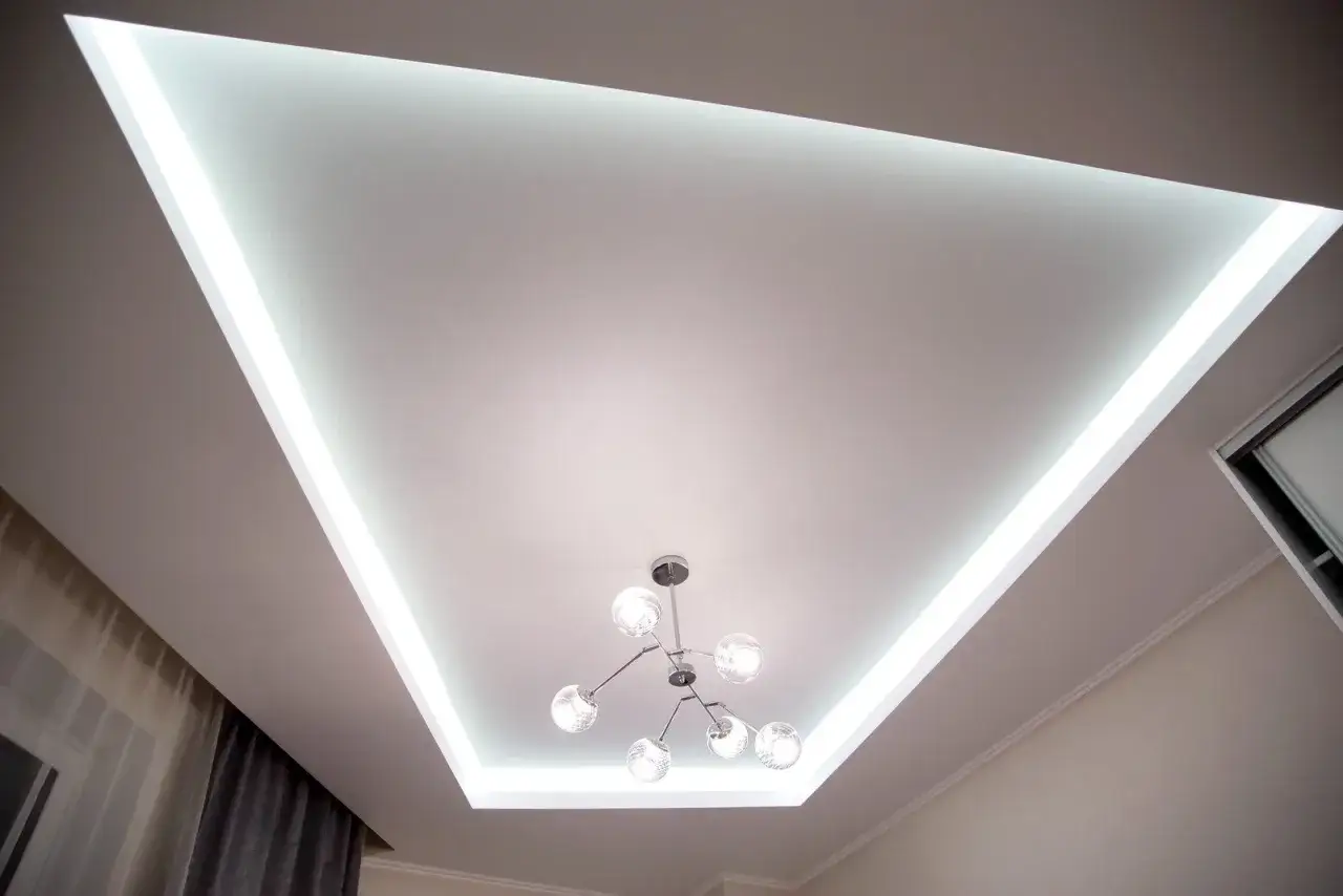 Nowoczesny sufit napinany z podświetleniem LED tworzy trójkątny kształt, a pośrodku wisi designerska lampa.