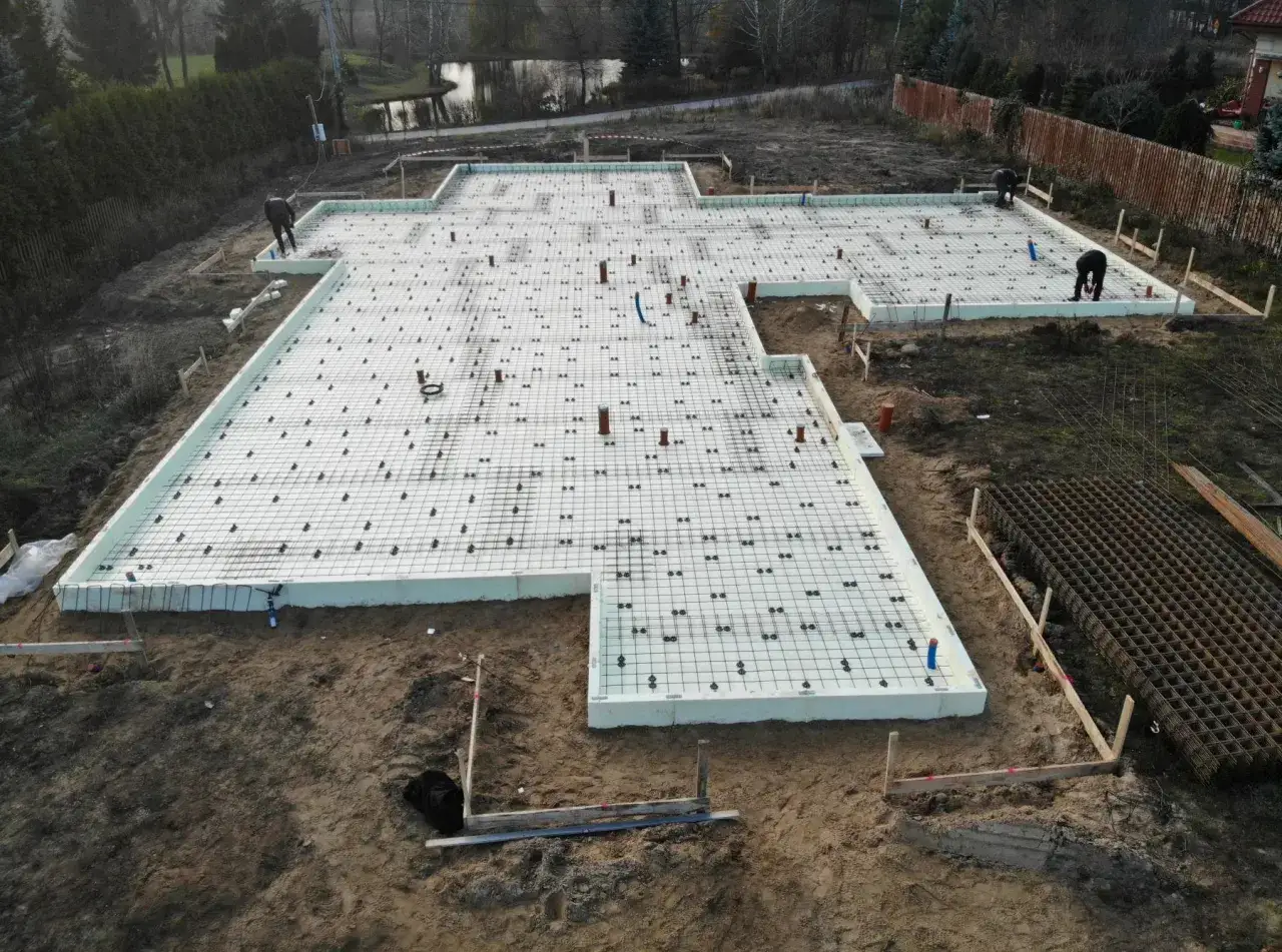 Budowa fundamentów pod dom. Widać zbrojenie i styropian. W tle staw i drzewa. Powstaje taras na słupach betonowych.